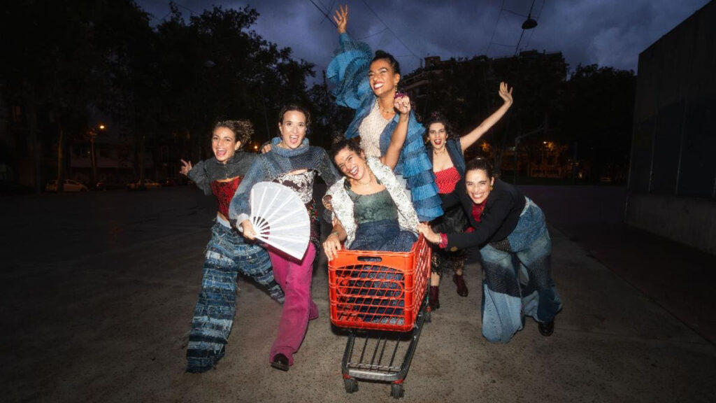 Six cheerful performers in colorful patched outfits pose around a red shopping cart on a nighttime street, arms raised.