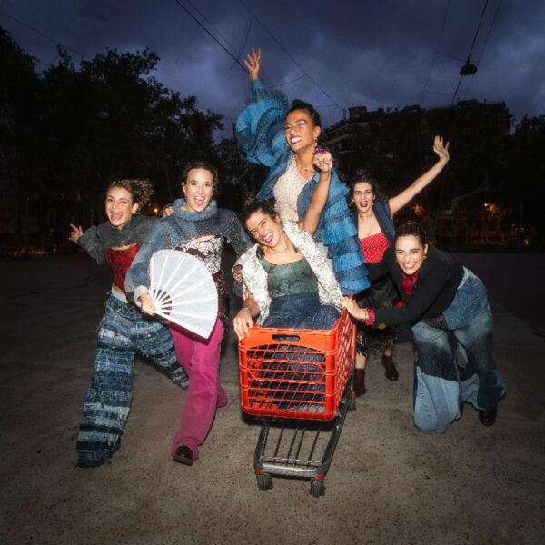 Six cheerful performers in colorful patched outfits pose around a red shopping cart on a nighttime street, arms raised.