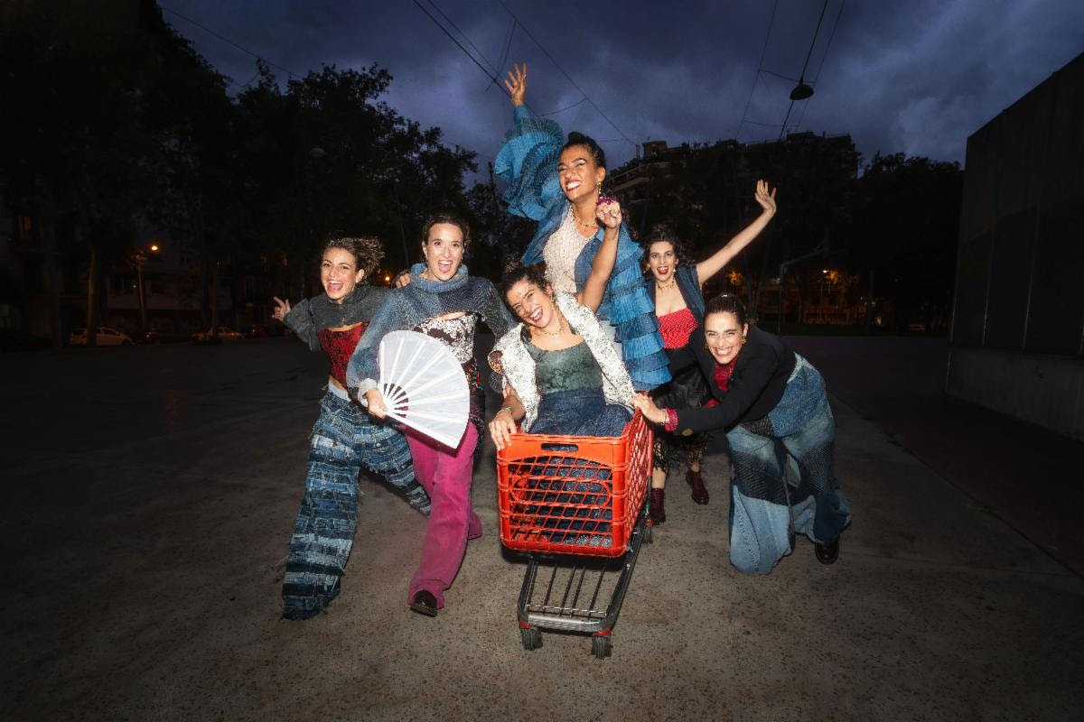 Six cheerful performers in colorful patched outfits pose around a red shopping cart on a nighttime street, arms raised.
