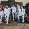 Five people in light-blue suits with colorful ties posing among stacked tires in a junkyard.