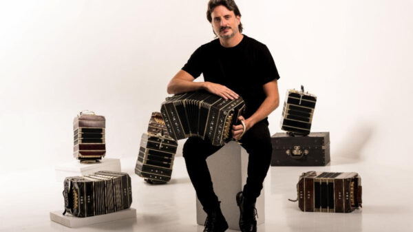 Male musician in black shirt seated on a block, playing a large accordion with several accordions arranged around him in a white studio.