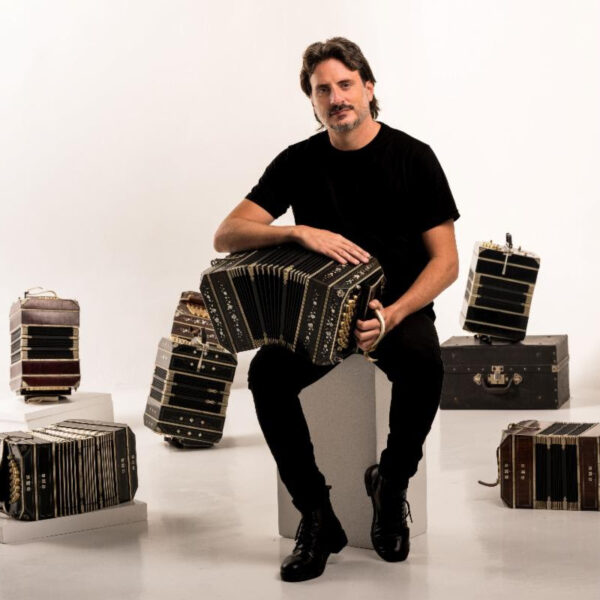 Male musician in black shirt seated on a block, playing a large accordion with several accordions arranged around him in a white studio.