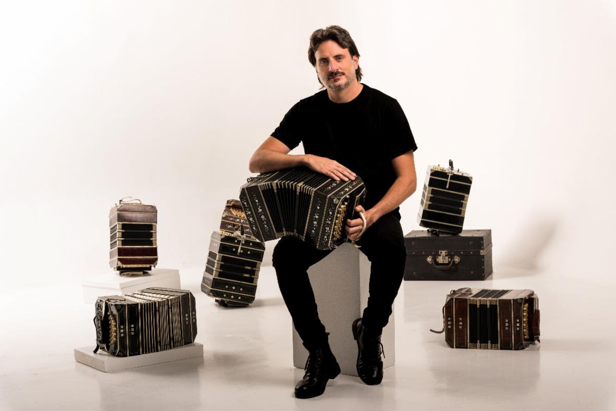Male musician in black shirt seated on a block, playing a large accordion with several accordions arranged around him in a white studio.