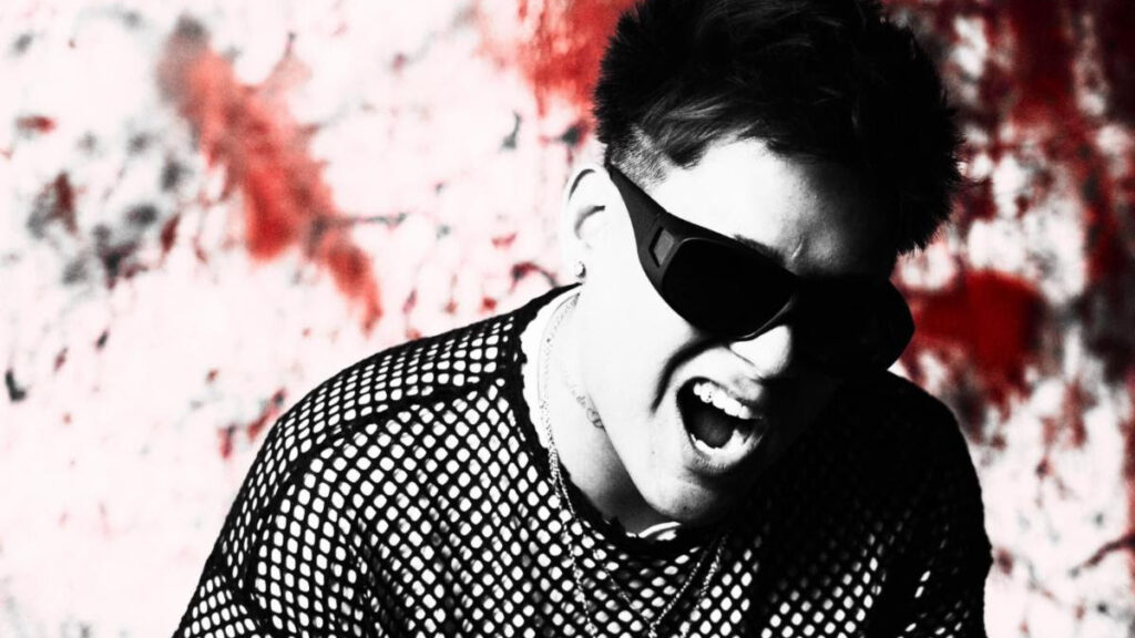 Man wearing dark sunglasses and a mesh shirt, shouting or singing against a red graffiti wall background, high-energy pose.