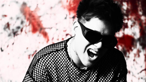 Man wearing dark sunglasses and a mesh shirt, shouting or singing against a red graffiti wall background, high-energy pose.