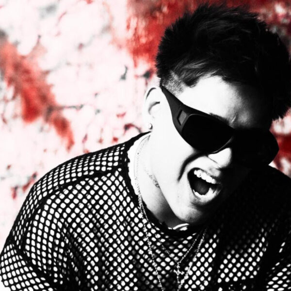 Man wearing dark sunglasses and a mesh shirt, shouting or singing against a red graffiti wall background, high-energy pose.
