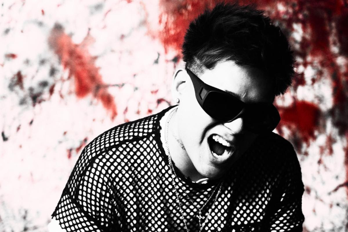 Man wearing dark sunglasses and a mesh shirt, shouting or singing against a red graffiti wall background, high-energy pose.