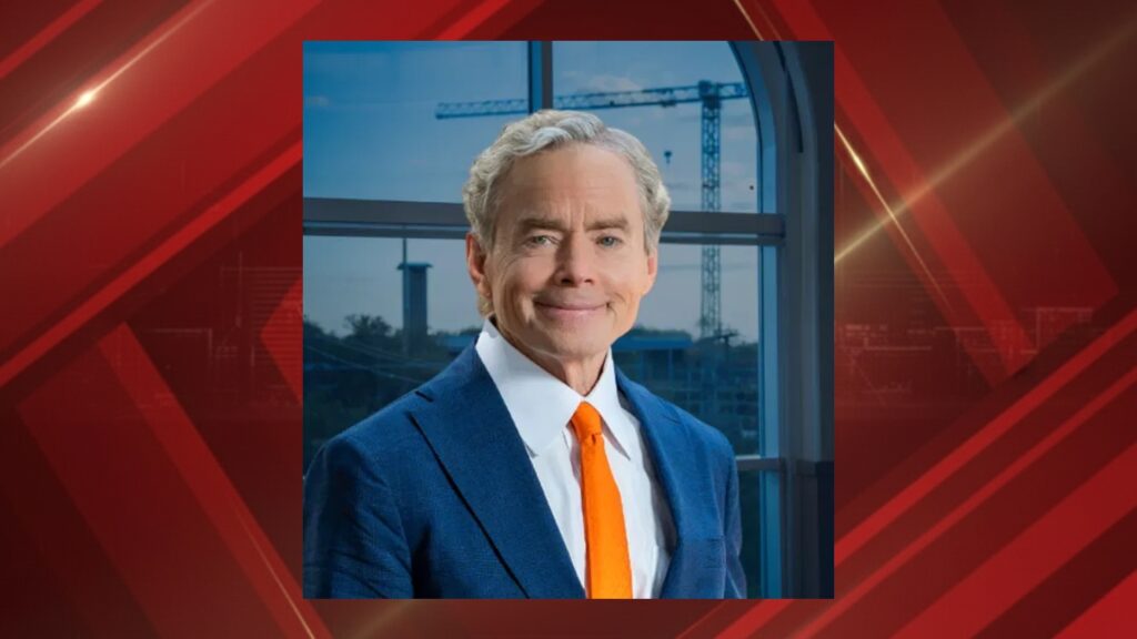 Portrait of a smiling man in a blue suit and orange tie standing by a window with construction cranes outside.