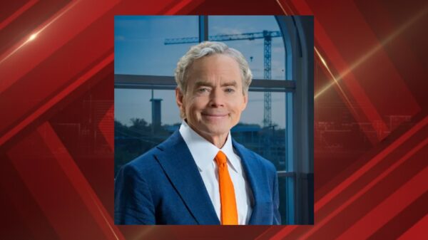 Portrait of a smiling man in a blue suit and orange tie standing by a window with construction cranes outside.