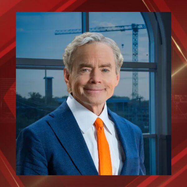 Portrait of a smiling man in a blue suit and orange tie standing by a window with construction cranes outside.