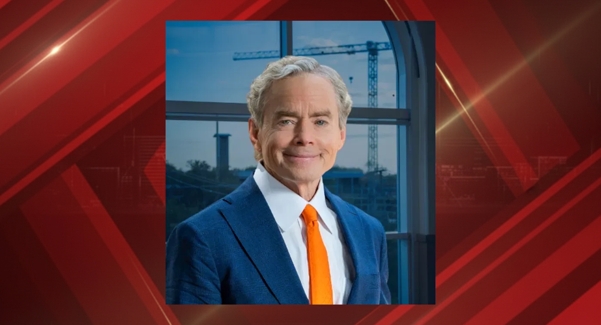 Portrait of a smiling man in a blue suit and orange tie standing by a window with construction cranes outside.
