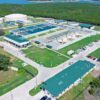 Aerial view of a large water treatment facility with circular clarifier tanks, rectangular filtration basins, green-roofed buildings, and a parking lot set among trees by a blue river or lake edge.