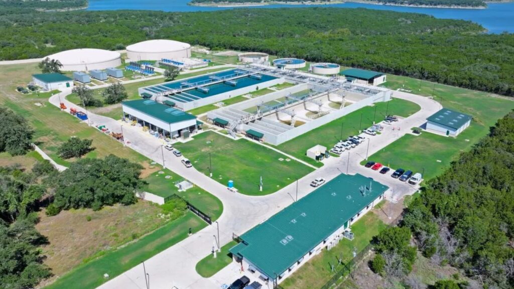 Aerial view of a large water treatment facility with circular clarifier tanks, rectangular filtration basins, green-roofed buildings, and a parking lot set among trees by a blue river or lake edge.