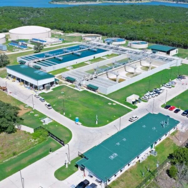 Aerial view of a large water treatment facility with circular clarifier tanks, rectangular filtration basins, green-roofed buildings, and a parking lot set among trees by a blue river or lake edge.