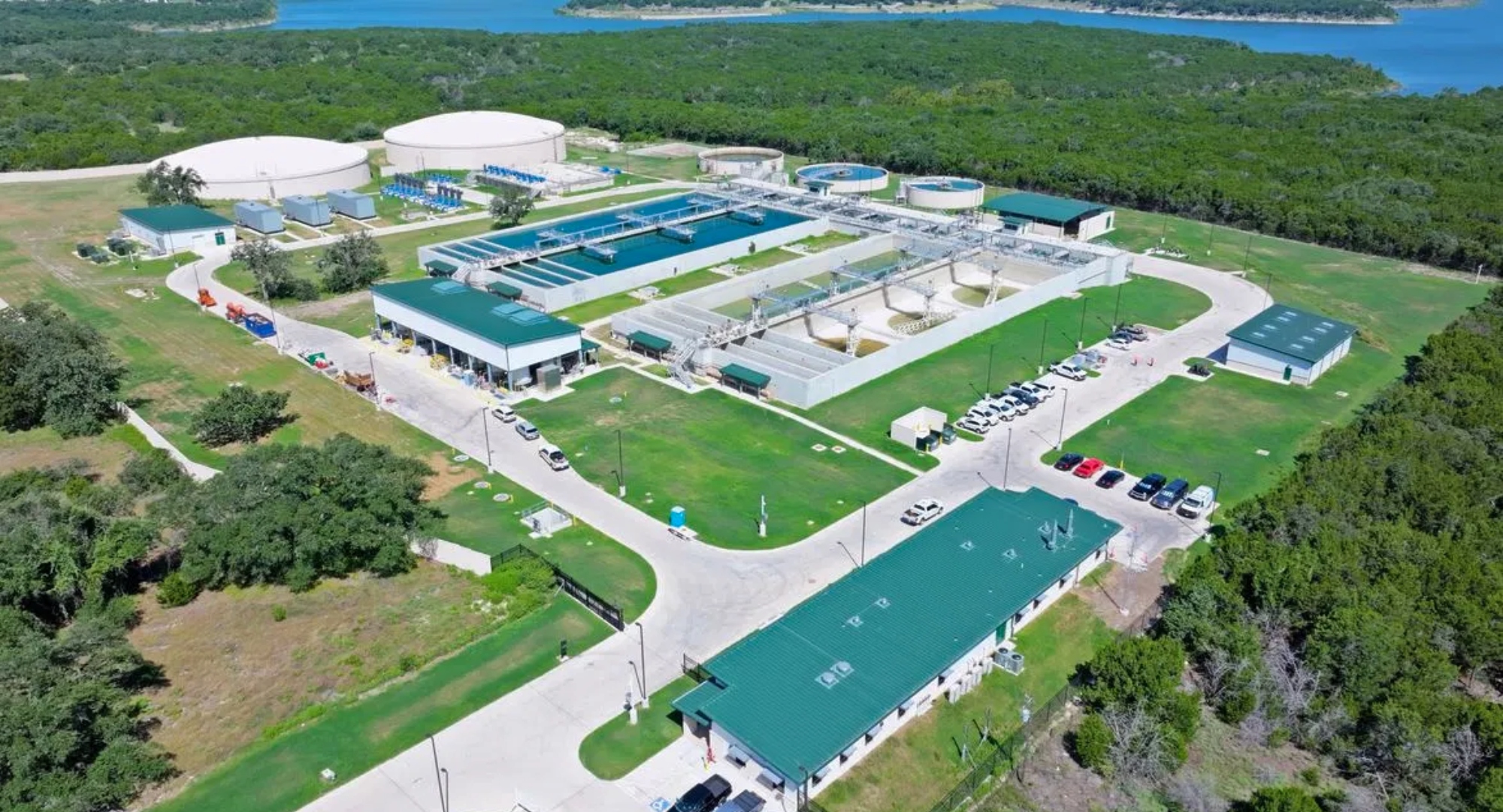 Aerial view of a large water treatment facility with circular clarifier tanks, rectangular filtration basins, green-roofed buildings, and a parking lot set among trees by a blue river or lake edge.
