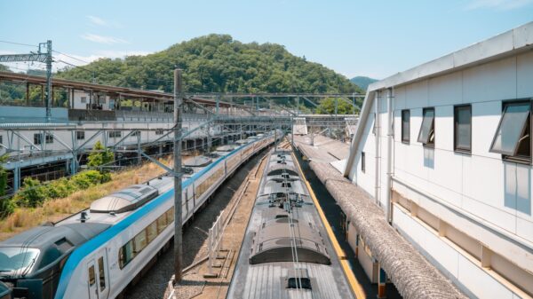 tren - Austin Latino Two high‑speed trains at a station platform with overhead electrical wires and a white building, hills in the background.",