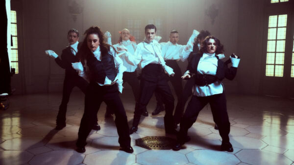 A group of dancers in white shirts and black pants performing a synchronized routine in a lit studio courtyard setting.