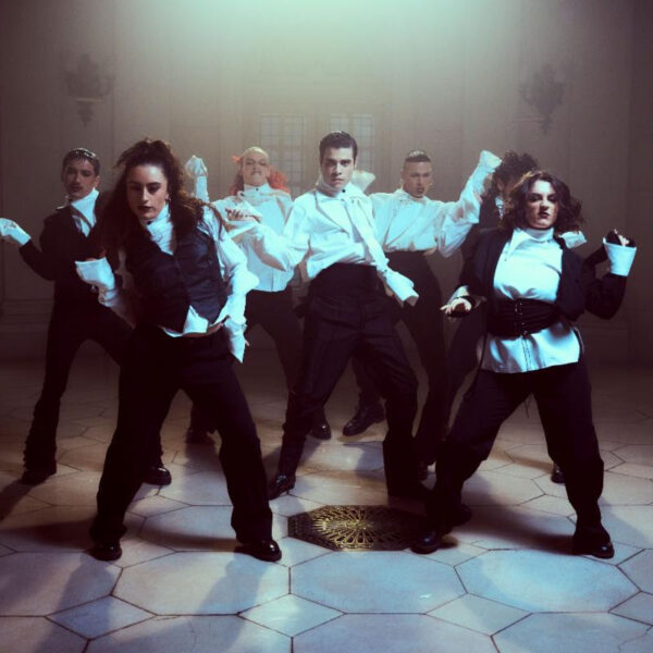 A group of dancers in white shirts and black pants performing a synchronized routine in a lit studio courtyard setting.