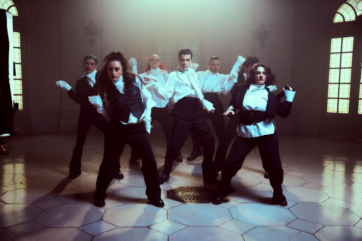A group of dancers in white shirts and black pants performing a synchronized routine in a lit studio courtyard setting.