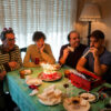 Four friends sit around a table with a lit birthday cake and candles, celebrating together at home.