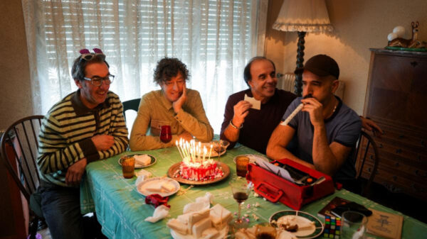 Four friends sit around a table with a lit birthday cake and candles, celebrating together at home.