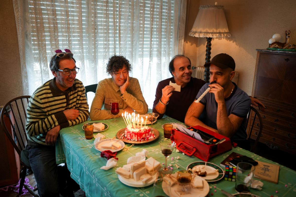 Four friends sit around a table with a lit birthday cake and candles, celebrating together at home.
