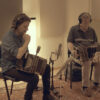 Two men sit in a recording studio playing accordions with headphones and microphones nearby.