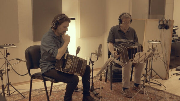 Two men sit in a recording studio playing accordions with headphones and microphones nearby.