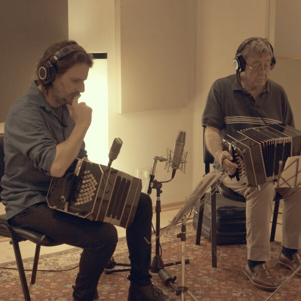 Two men sit in a recording studio playing accordions with headphones and microphones nearby.