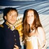 Two people posing for a photo at night: a man in a dark navy military-style jacket with gold trim and a woman in a cream corset dress stand before a blue-and-white backdrop.