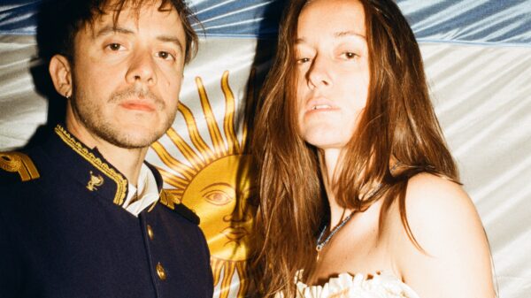 Two people posing for a photo at night: a man in a dark navy military-style jacket with gold trim and a woman in a cream corset dress stand before a blue-and-white backdrop.