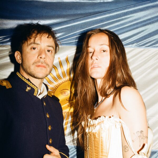 Two people posing for a photo at night: a man in a dark navy military-style jacket with gold trim and a woman in a cream corset dress stand before a blue-and-white backdrop.