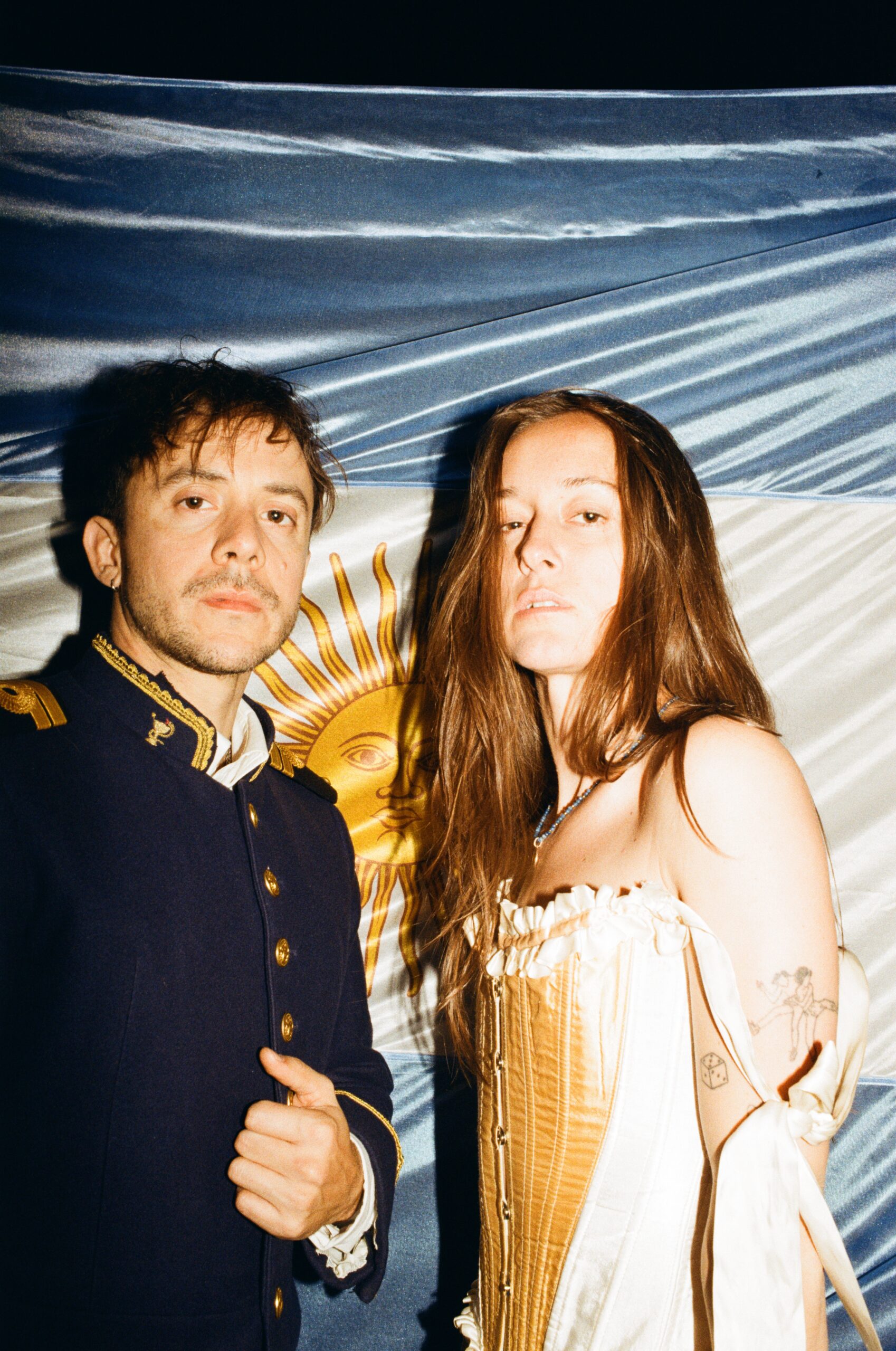 Two people posing for a photo at night: a man in a dark navy military-style jacket with gold trim and a woman in a cream corset dress stand before a blue-and-white backdrop.
