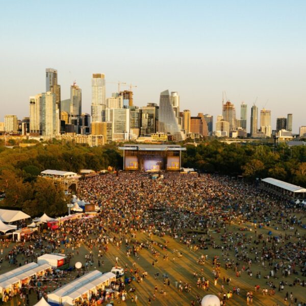 Large outdoor concert in a park with a packed crowd and a stage, city skyline in the background at dusk.