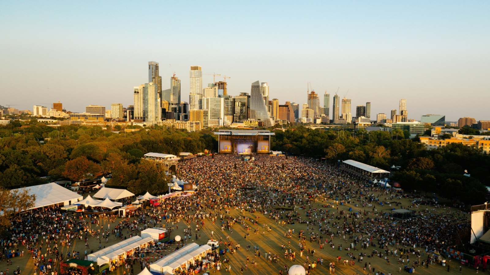 Large outdoor concert in a park with a packed crowd and a stage, city skyline in the background at dusk.
