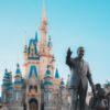 Statues of a man in a suit with one hand raised beside a Mickey Mouse statue in front of a blue-roofed castle at a Disney park