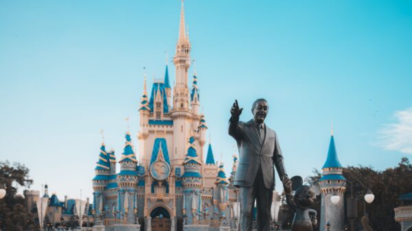 Statues of a man in a suit with one hand raised beside a Mickey Mouse statue in front of a blue-roofed castle at a Disney park