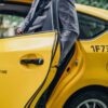 Yellow taxi with a rear door ajar as a person in a gray suit steps into the back seat; markings show 'Metered Fare' and 'Flat Fare JFK' on the door.