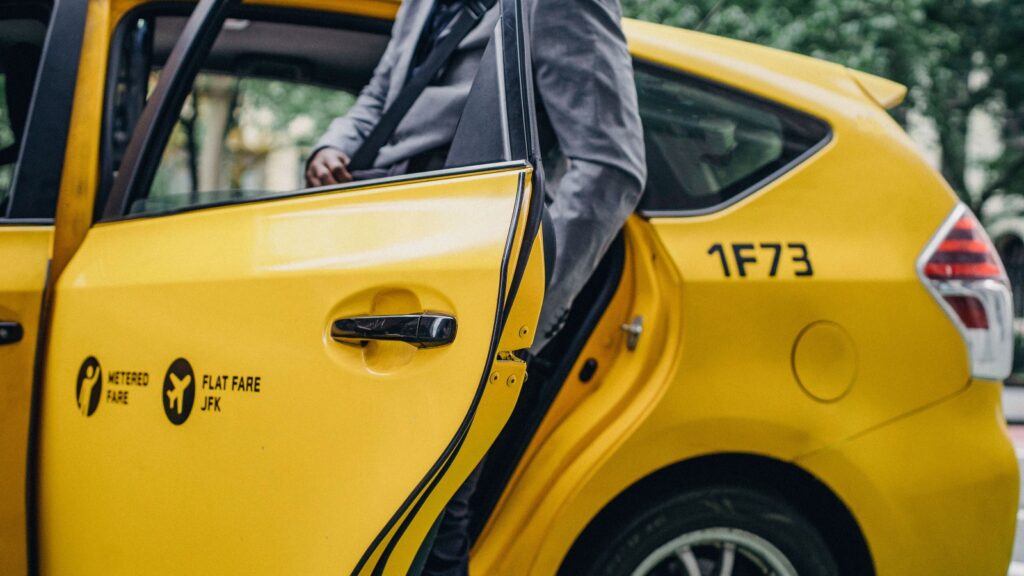 Yellow taxi with a rear door ajar as a person in a gray suit steps into the back seat; markings show 'Metered Fare' and 'Flat Fare JFK' on the door.