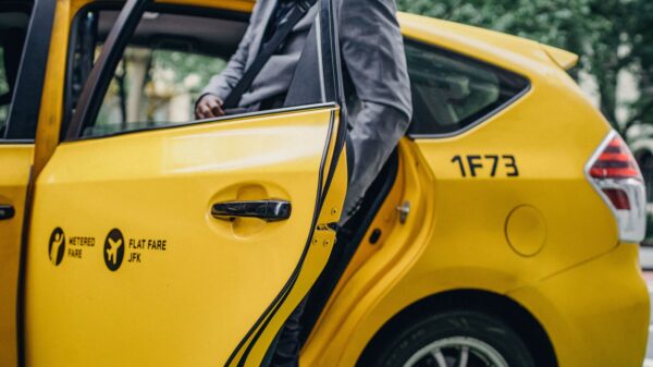 Yellow taxi with a rear door ajar as a person in a gray suit steps into the back seat; markings show 'Metered Fare' and 'Flat Fare JFK' on the door.