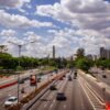 Busy urban highway with multiple lanes and cars, flanked by trees, under a blue sky with fluffy clouds.