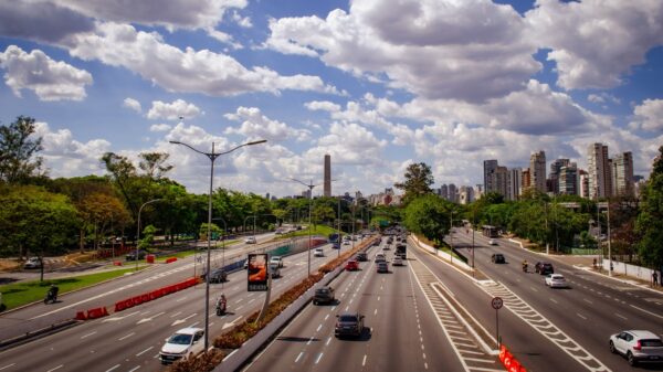 Busy urban highway with multiple lanes and cars, flanked by trees, under a blue sky with fluffy clouds.