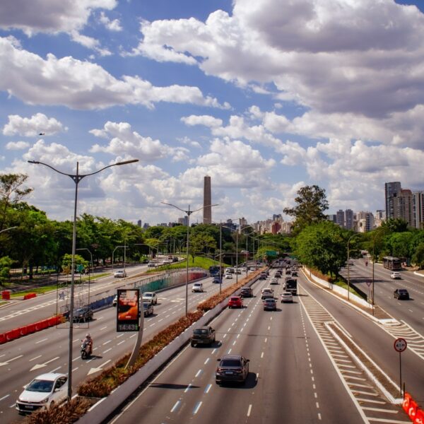 Busy urban highway with multiple lanes and cars, flanked by trees, under a blue sky with fluffy clouds.