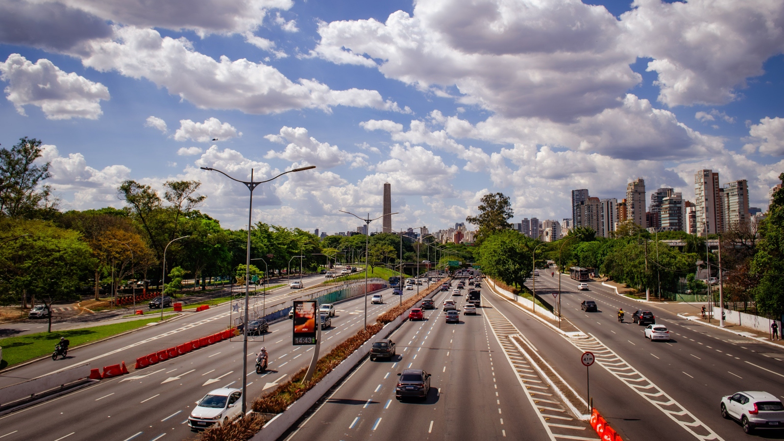 Busy urban highway with multiple lanes and cars, flanked by trees, under a blue sky with fluffy clouds.