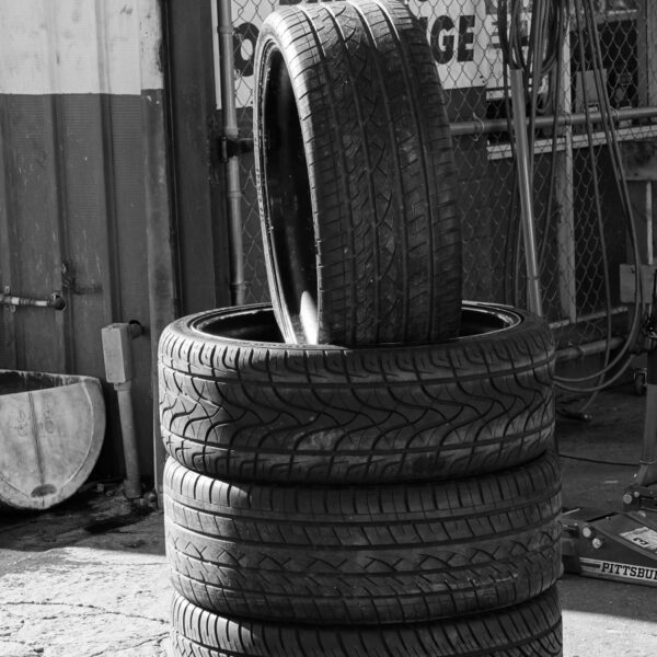 Stack of four car tires outside a repair shop, with one tire leaned upright on top; jack stands and tools nearby.