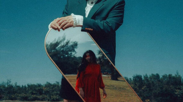 Man in a teal suit outdoors holds a tilted mirror reflecting a woman in a red dress in a grassy field.