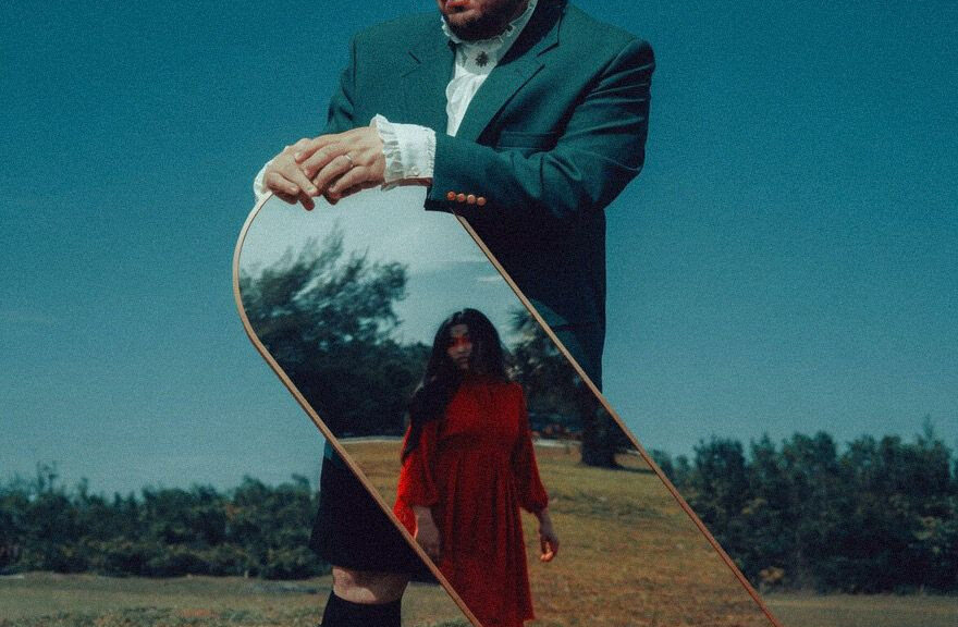 Man in a teal suit outdoors holds a tilted mirror reflecting a woman in a red dress in a grassy field.