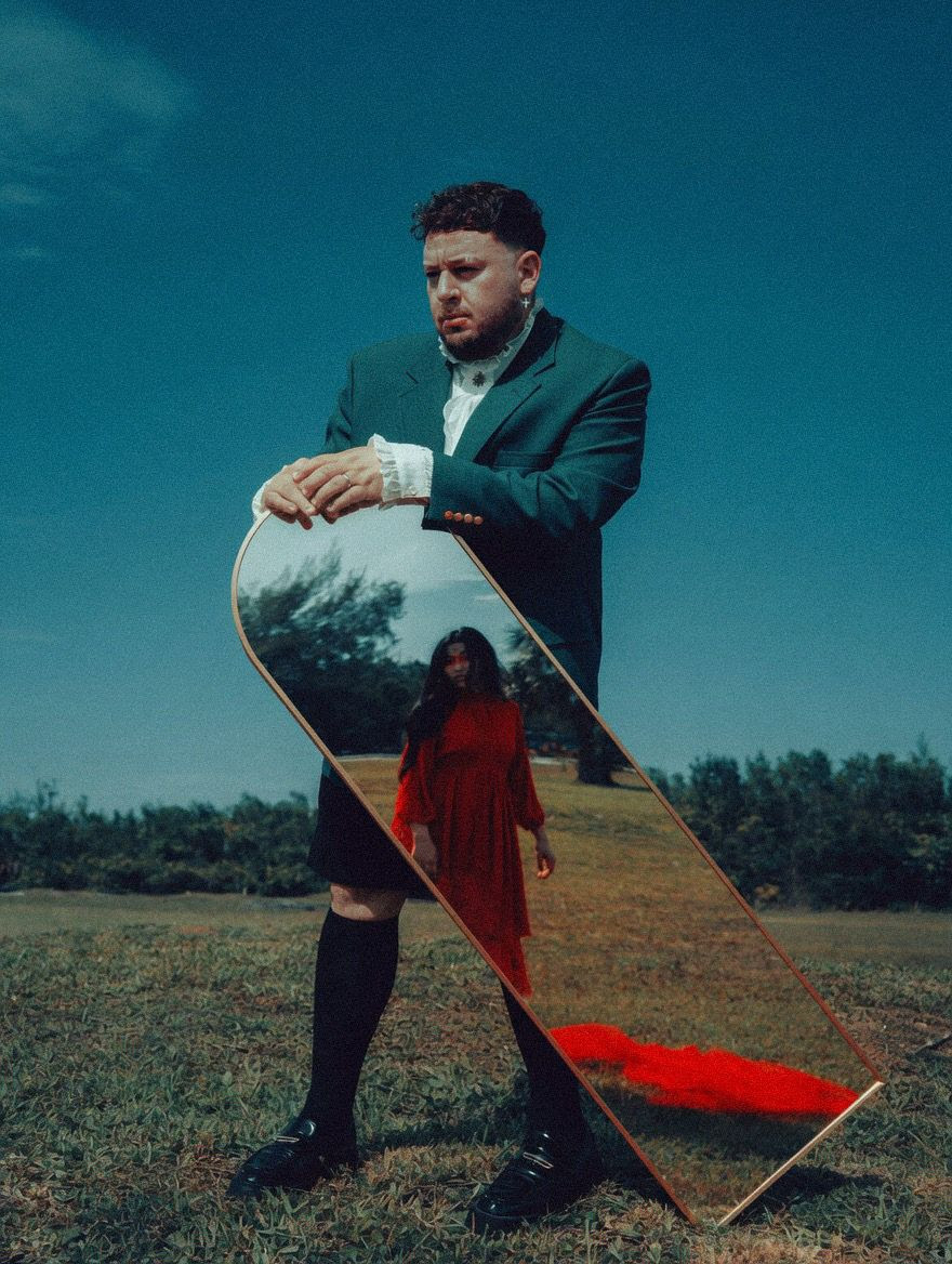 Man in a teal suit outdoors holds a tilted mirror reflecting a woman in a red dress in a grassy field.