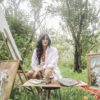 Woman artist seated outdoors with paints and brushes at a table, surrounded by framed artworks on the ground and easel art in progress.
