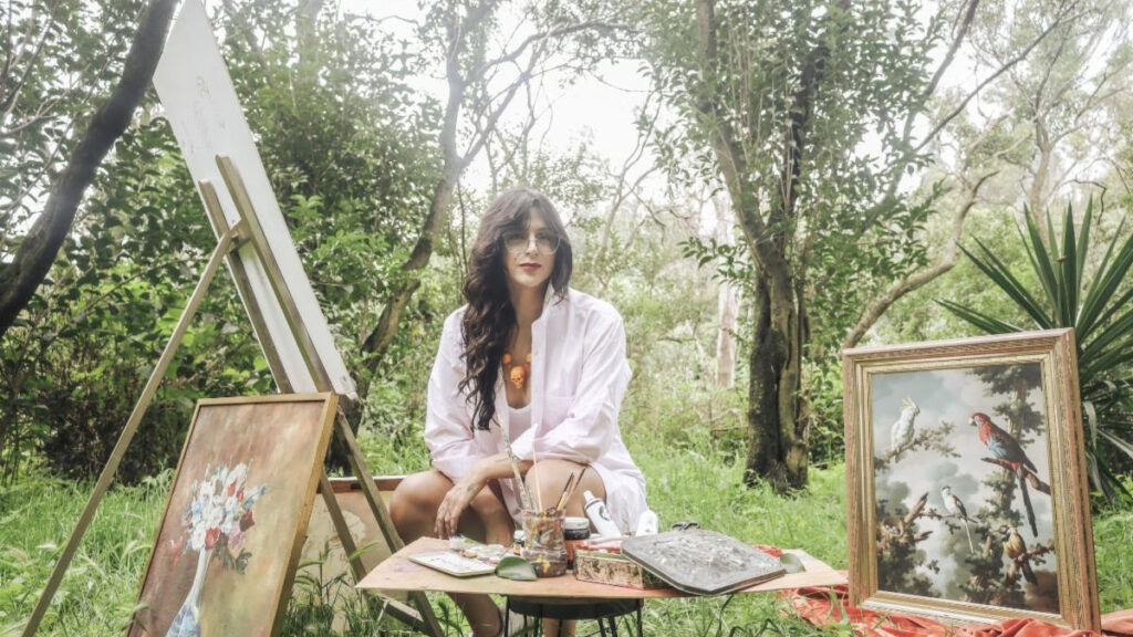 Woman artist seated outdoors with paints and brushes at a table, surrounded by framed artworks on the ground and easel art in progress.